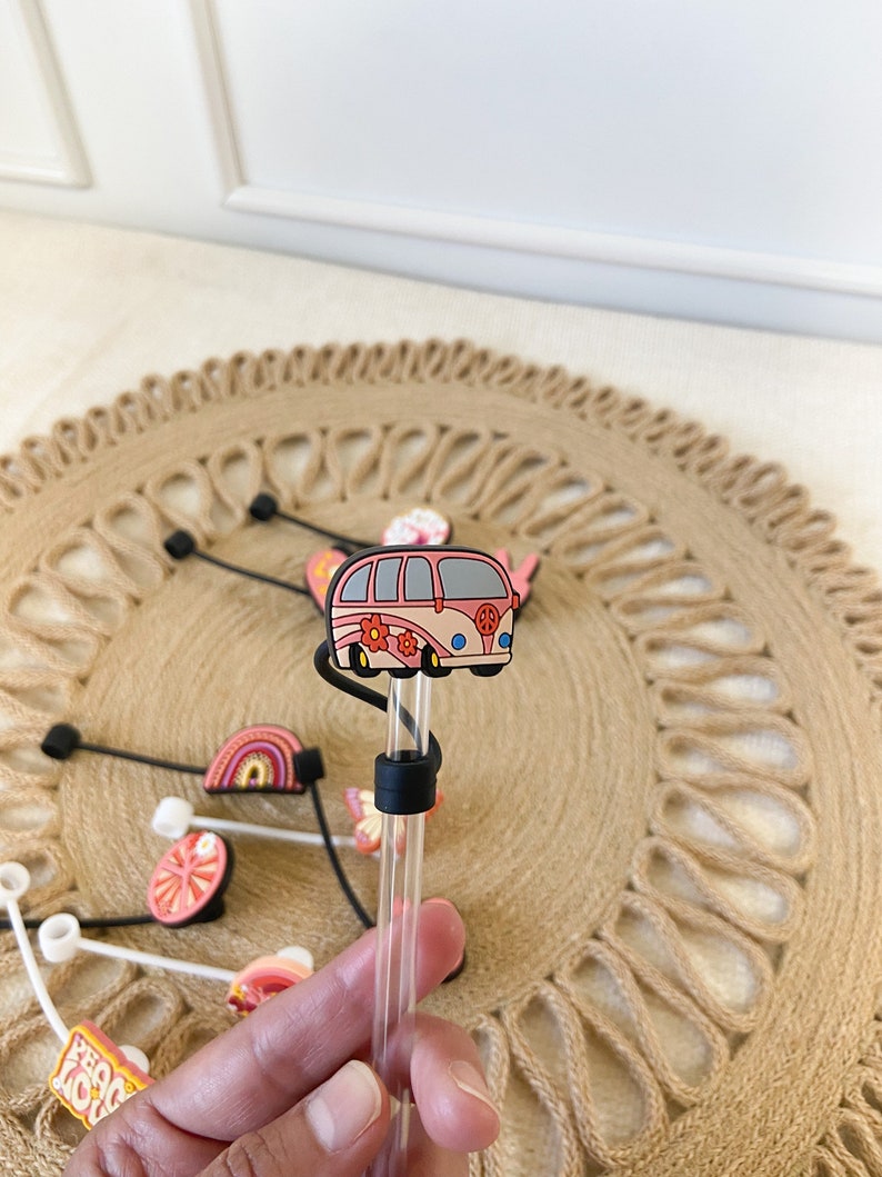 May include: A clear straw with a black silicone ring and a decorative topper featuring a pink and peach-colored retro van with flower accents. Other straw toppers with rainbows, butterflies, and fruit are also visible.