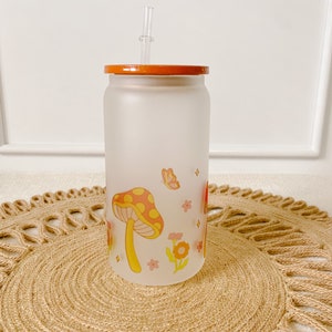May include: A frosted glass tumbler with a wooden lid and a clear plastic straw. The tumbler features a whimsical design of red and yellow mushrooms, pink flowers, and butterflies on a white background.