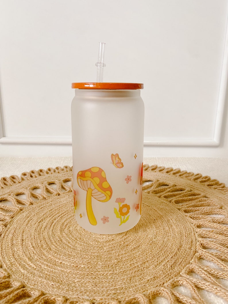 May include: A frosted glass tumbler with a wooden lid and a clear plastic straw. The tumbler features a whimsical design of red and yellow mushrooms, pink flowers, and butterflies on a white background.