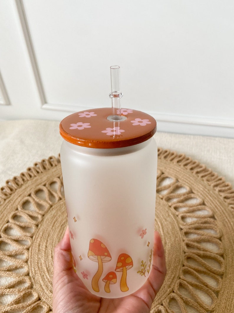 May include: A frosted glass water bottle with a brown lid and pink flowers. The bottle has a mushroom design with red and yellow accents. A clear plastic straw is inserted through the lid.