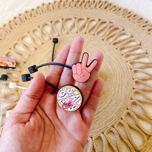 May include: A hand holding two black hair ties with decorative charms. One charm is a pink peace sign, and the other is a round charm with the words "Stay Groovy" in pink and white. The background is a woven, circular placemat.