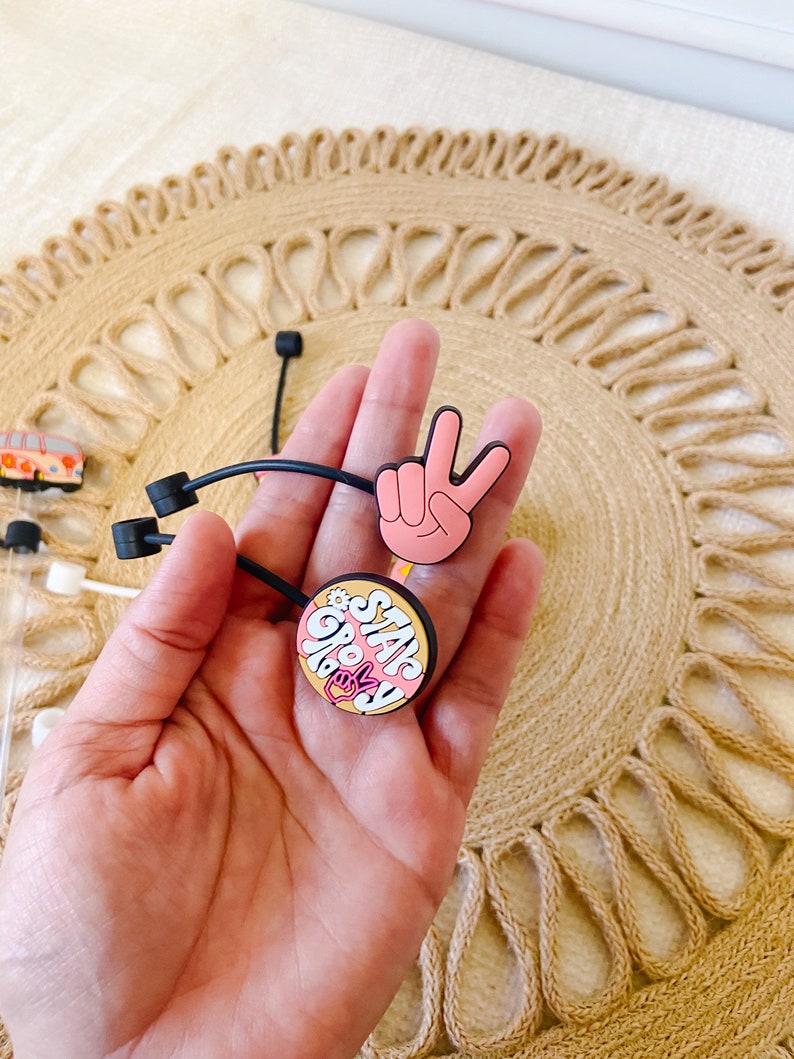 May include: A hand holding two black hair ties with decorative charms. One charm is a pink peace sign, and the other is a round charm with the words "Stay Groovy" in pink and white. The background is a woven, circular placemat.