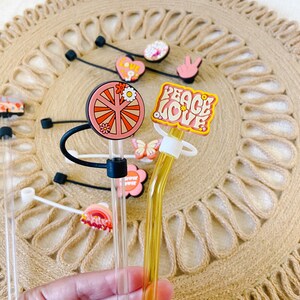 May include: A collection of clear glass straws with decorative toppers. The toppers feature various designs, including a peace sign, a flower, a heart, and the words "Peace Love". The straws are displayed on a woven mat.