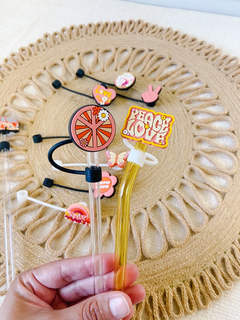 May include: A collection of clear glass straws with decorative toppers. The toppers feature various designs, including a peace sign, a flower, a heart, and the words "Peace Love". The straws are displayed on a woven mat.