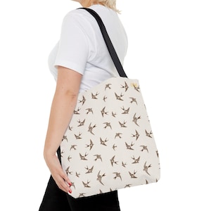 Peut inclure: Un sac fourre-tout en toile blanche avec des sangles noires et un motif répété d'hirondelles brunes. Le sac a une fermeture à bouton dorée.