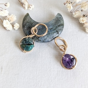 May include: Two gold-toned wire wrapped charms with gemstones. The charm on the left features a green stone, and the charm on the right features a purple stone. Both charms are on a gray crescent moon-shaped stone.
