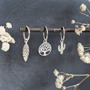 May include: Three silver hoop earrings with different charms: a feather, a tree, and a cactus. The earrings are hanging from a black bar.