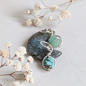 May include: A silver wire wrapped charm with a turquoise stone and two green stones. The charm is sitting on a gray moon-shaped stone.