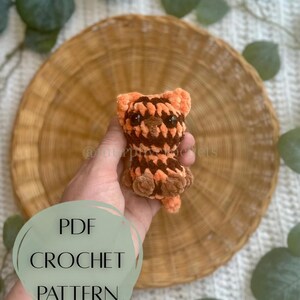 Op de afbeelding: Een kleine, handgemaakte gehaakte tijgerwelp in bruin- en oranjetinten. De welp wordt in een hand gehouden, met een ronde, geweven placemat op de achtergrond. De tekst "PDF CROCHET PATTERN" staat over de afbeelding.