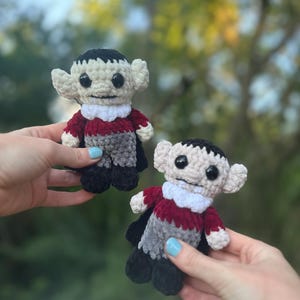 May include: Two crocheted plush vampire toys with black hair, red and gray clothing, and white collars. The toys have black eyes and white faces.