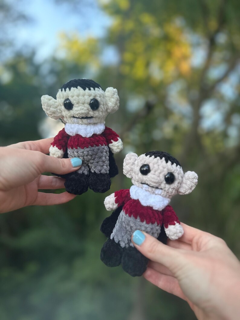 May include: Two crocheted plush vampire toys with black hair, red and gray clothing, and white collars. The toys have black eyes and white faces.