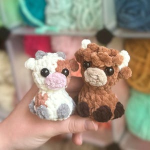 May include: Two crocheted cow figurines. One is white with brown and gray spots, a pink nose, and black eyes. The other is brown with white accents and black eyes. Both are small and plush, held in a hand.