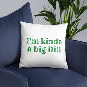 May include: White square pillow with the text "I'm kinda a big Dill" in green, decorative font. The pillow is placed on a dark blue armchair, with green plants in the background.