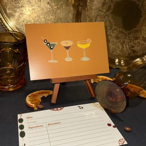 May include: A brown card on a wooden easel displays three martini illustrations with olives, coffee beans, and a lemon twist. A recipe card with lines for title, servings, ingredients, and directions is in front.