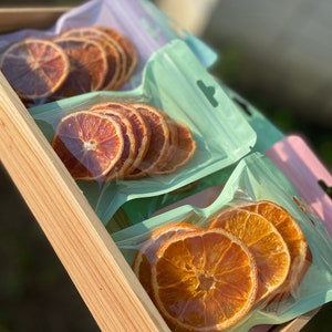 May include: Three clear plastic bags filled with dried orange slices. The bags are arranged in a row on a wooden surface. The orange slices are a vibrant orange color and are stacked on top of each other.