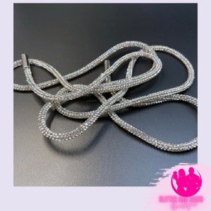 May include: A silver-colored, rhinestone-studded drawstring cord. The cord is made of a flexible material and is covered in small, sparkling rhinestones. The cord is looped and intertwined, creating a decorative effect. The cord has metal tips.