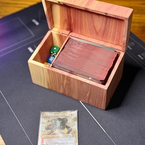 May include: A wooden card storage box with a hinged lid, open to reveal a stack of red-edged cards. The box also contains dice. A card with the text "Quintorius, History Chaser" is in front of the box.