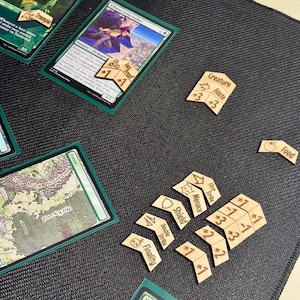 May include: A collection of Magic: The Gathering cards and wooden tokens on a black surface. The cards feature green borders and fantasy art. Wooden tokens include labels like "Creature," "Haste," and "Food," along with numerical values.