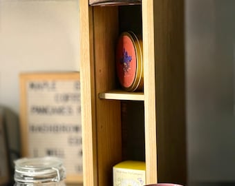 May include: A wooden tea bag and tea tin storage cabinet with a sliding top. A decorative mug with a floral design sits on the counter. A jar of dried orange slices is in the background.