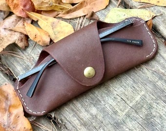 May include: A brown leather glasses case with a snap closure, holding a pair of black-framed glasses. The case has white stitching around the edges and a gold-colored snap. The glasses are partially visible, with the brand name "TED BAKER" on the arm.