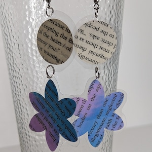 May include: A pair of earrings with a flower design. The flowers are made of clear plastic with a printed design of text. The text is from a book and is in a light blue and purple color scheme. The earrings are hanging from silver hooks.