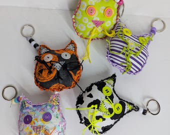 May include: Five colorful fabric cat keychains with different patterns and button eyes. The keychains are made of different fabrics, including black and white, green and white, purple and white, orange and black, and pink and white.