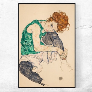 May include: A framed print of an artwork depicting a seated figure. The figure has red hair and is wearing a green top, white shorts, and black leggings. The artwork is a line drawing with watercolor accents.