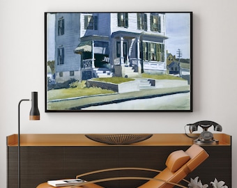 May include: Framed print of a house painting. The artwork depicts a two-story house with a porch and green shutters. The painting is in a black frame and hangs above a modern brown cabinet with a leather chaise lounge.