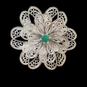 Puede incluir: Un broche de flor de filigrana de plata con una piedra preciosa verde en el centro.