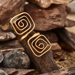 May include: Two gold rings with a spiral design. The rings are stacked on top of each other and are sitting on a piece of driftwood.