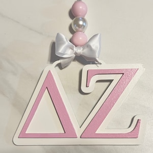 May include: A pink and white wooden sign with the letters "ΔZ" in a layered design. The sign has a white bow and a pink and white beaded garland attached to the top.