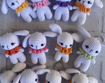 May include: A collection of white crochet bunny keychains with various colored ruffled collars. The bunnies have black eyes and a 'Y' shaped nose. The collars are in shades of yellow, pink, purple, orange, and green. The keychains are displayed against a light gray background.