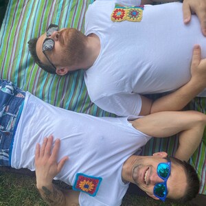 May include: Two people wearing white t-shirts with colorful crocheted squares on the front. The person on the left is wearing sunglasses and lying on a striped blanket. The person on the right is wearing blue sunglasses and lying on grass.