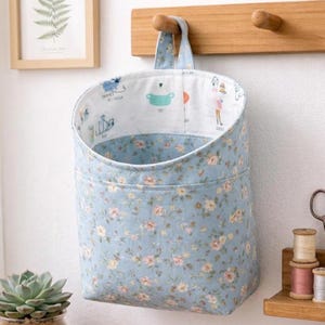 May include: A hanging fabric storage basket with a light blue exterior featuring a small floral pattern. The interior lining is white with various small colourful illustrations and text. The basket has a fabric loop for hanging on a wall hook.