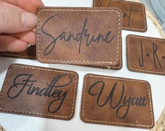 May include: Five brown leather place cards with black cursive lettering. The names on the cards are Sandrine, Findlay, Wyatt, J-R, and a name that is partially obscured.