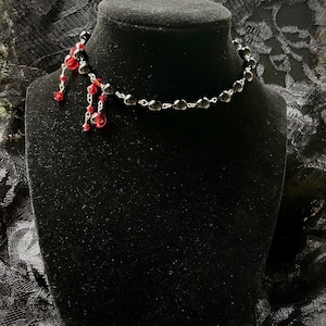 Puede incluir: Un collar gargantilla de cuentas negras con cuentas rojas colgantes. El collar se exhibe sobre un busto de maniquí negro sobre un fondo oscuro. La gargantilla presenta pequeñas cuentas negras redondas y una cadena plateada.