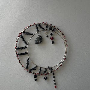 May include: A silver-toned wire crescent moon pendant necklace with black and red beads. The necklace features a black stone pendant and dangling black and red beads. The design is set against a white background.