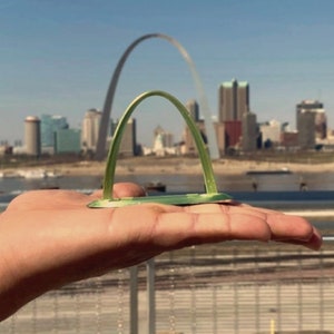May include: A miniature green arch model, held in a hand, with the Gateway Arch in the background. The model is a small, detailed representation of the iconic landmark, set against a cityscape and a clear blue sky.