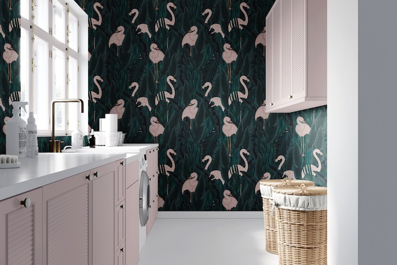 May include: A laundry room with a flamingo-patterned wallpaper in shades of green and pink. The room features pink cabinets, a white countertop, a washing machine, and two wicker laundry baskets. A window and a gold faucet are also visible.