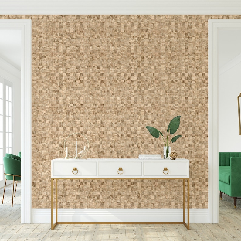 May include: A white console table with gold metal legs and three drawers. The table is placed in front of a wall with a brown textured wallpaper. A green plant and a decorative object are on top of the table.