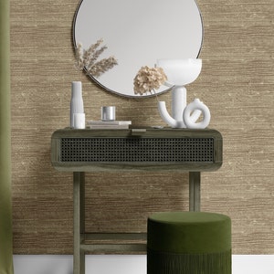 May include: A wooden console table with a woven rattan drawer and a green velvet ottoman with a fringe trim. The table is decorated with a round mirror, a white vase with dried flowers, a white candle holder, and a book. The background is a brown textured wallpaper.