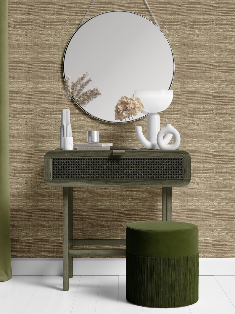 May include: A wooden console table with a woven rattan drawer and a green velvet ottoman with a fringe trim. The table is decorated with a round mirror, a white vase with dried flowers, a white candle holder, and a book. The background is a brown textured wallpaper.