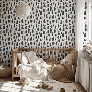 May include: A white crib with a black and white abstract patterned wallpaper behind it. The crib has a white blanket and a brown throw blanket on it. There are two white pillows and a stuffed animal on the crib.
