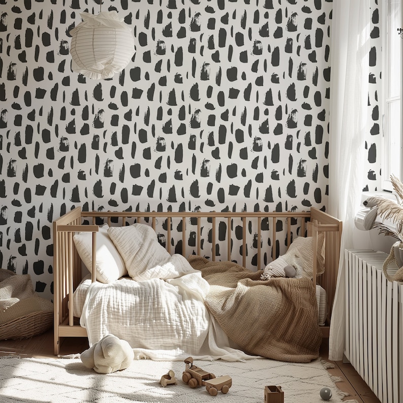 May include: A white crib with a black and white abstract patterned wallpaper behind it. The crib has a white blanket and a brown throw blanket on it. There are two white pillows and a stuffed animal on the crib.