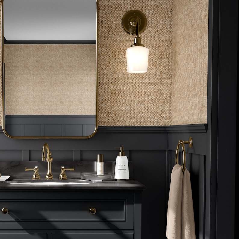 May include: A black bathroom vanity with a white porcelain sink and gold fixtures. The vanity has a single drawer and a towel rack on the right side. The walls are covered in a beige textured wallpaper. A gold light fixture with a frosted glass shade hangs above the mirror.