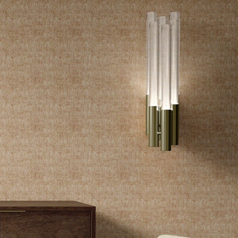 May include: A modern wall sconce with a brushed gold finish and clear glass tubes. The sconce is mounted on a beige textured wall.