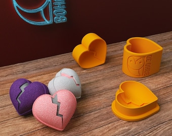 May include: Four broken heart-shaped molds, two are orange and two are pink, purple, and white. The molds are on a wooden surface. There are two whole heart-shaped molds, one is orange and one is yellow. The molds are all 3D printed and have the words "boho 3D" on them.