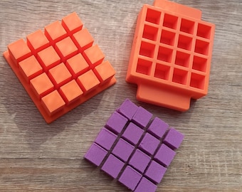 May include: Three silicone molds for making small square-shaped treats. The molds are orange and purple. The molds are on a wooden surface.