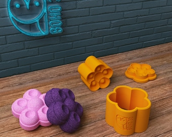 May include: Four silicone molds in the shape of a flower with six rounded petals. Two molds are purple, one is pink, and one is orange. The orange mold has the text "BOHO 3D" on the side. The molds are on a wooden surface.