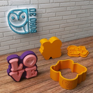 May include: Four cookie cutters in various shapes, including a heart, a cloud, and the word "LOVE". The cookie cutters are made of a bright yellow plastic and have a textured surface. The word "LOVE" is in pink and purple plastic. The cookie cutters are on a wooden surface.